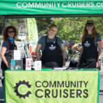 there smiling people standing under a green tent and in front of a table with a green banner that reads "Community Cruisers"