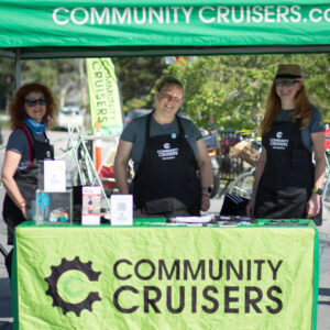 there smiling people standing under a green tent and in front of a table with a green banner that reads "Community Cruisers"